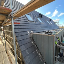 Dmytro Husak - Montaż elewacji drewnianej na budynku z dachem pokrytym czarną dachówką, widoczne rusztowania i narzędzia pracy.