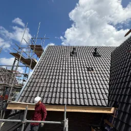 Prace dekarskie w Warszawie: Nowy dach z ciemnej dachówki ceramicznej, kominki wentylacyjne, widoczne rusztowania i dekarz w kasku podczas montażu.