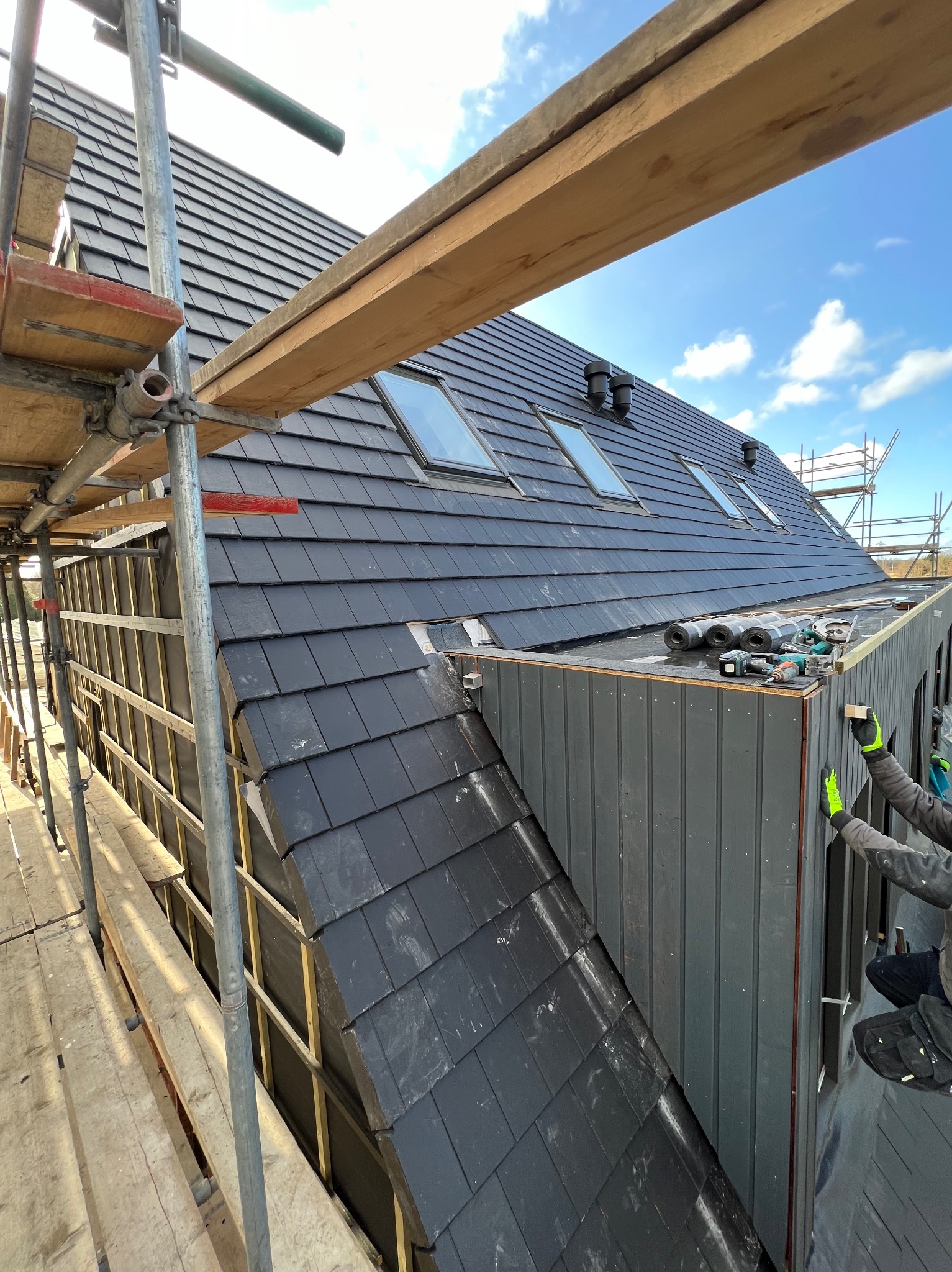 Montaż elewacji drewnianej na budynku z dachem pokrytym czarną dachówką, widoczne rusztowania i narzędzia pracy.