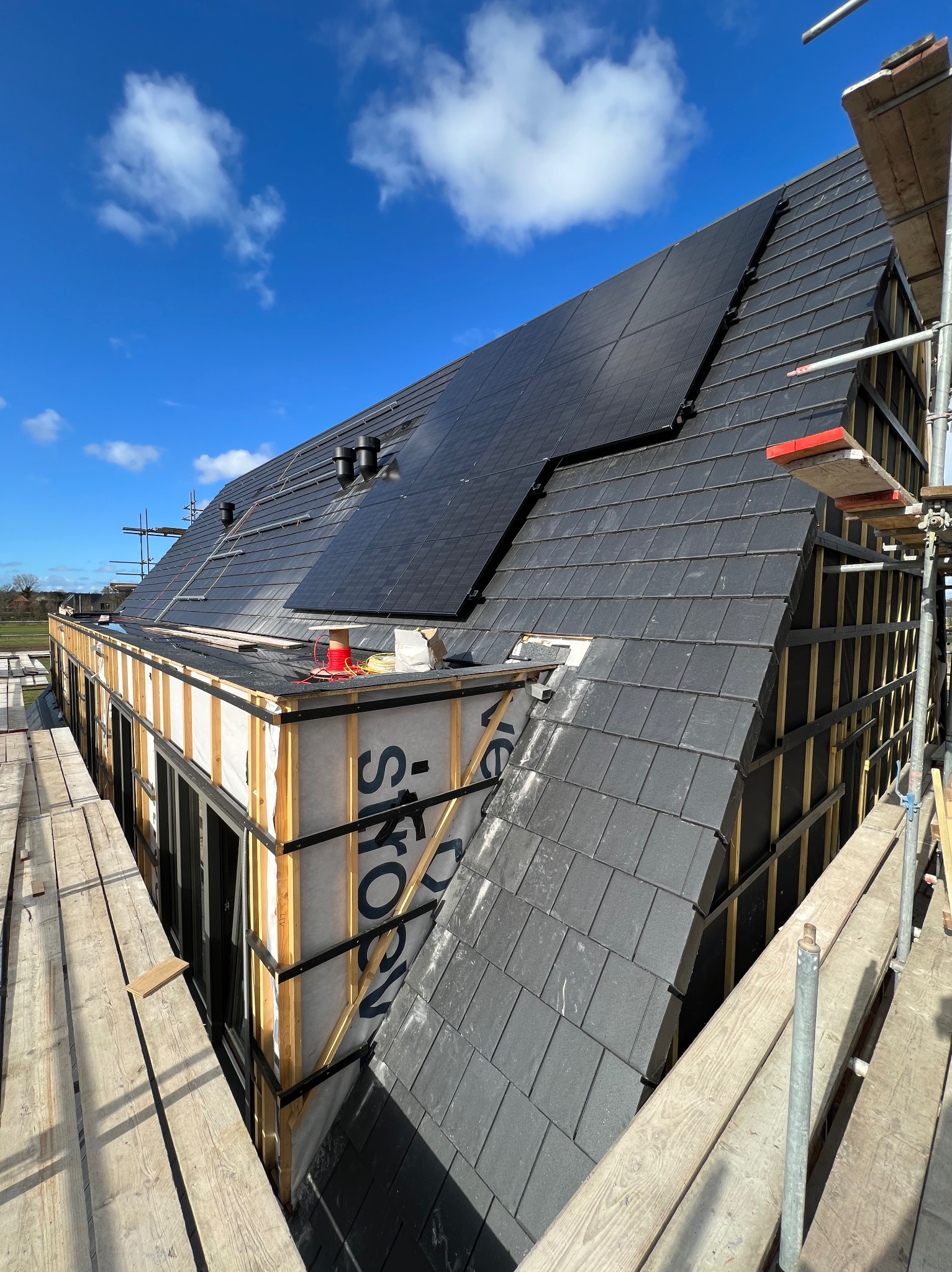 Nowy dach z ciemnej dachówki łupkowej i panelami słonecznymi w trakcie montażu, widok z rusztowania na tle błękitnego nieba z chmurami.