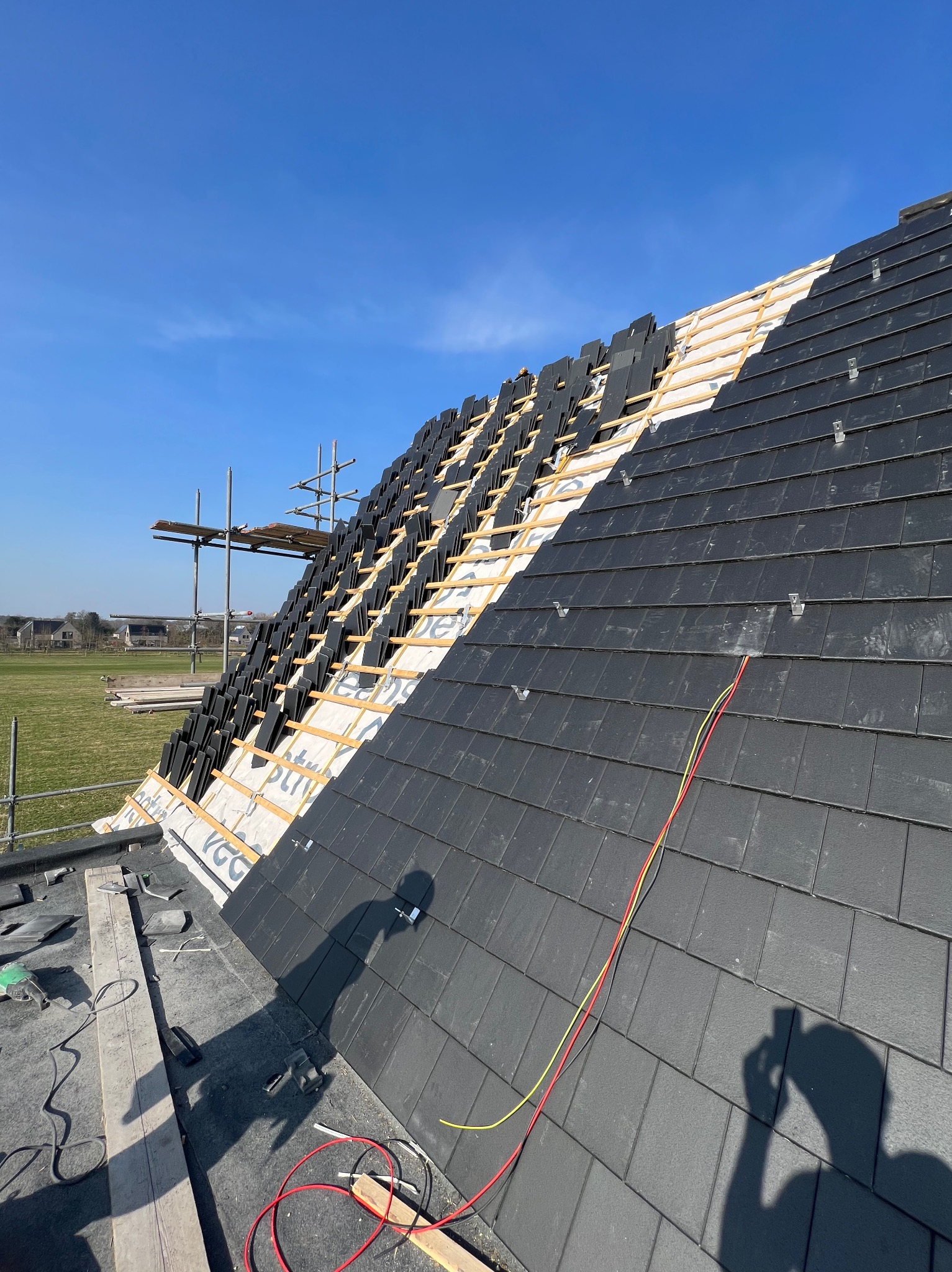 Częściowo pokryty dach z czarną dachówką, widoczna więźba i membrana, rusztowanie w tle, cień osoby robiącej zdjęcie w prawym dolnym rogu.
