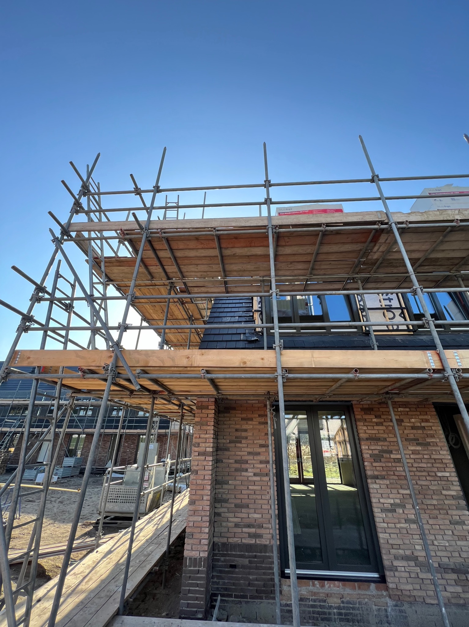 Budowa domu jednorodzinnego z cegły, widok z dołu na rusztowanie i fragment dachu pokrytego ciemną dachówką, okna z czarnymi ramami.