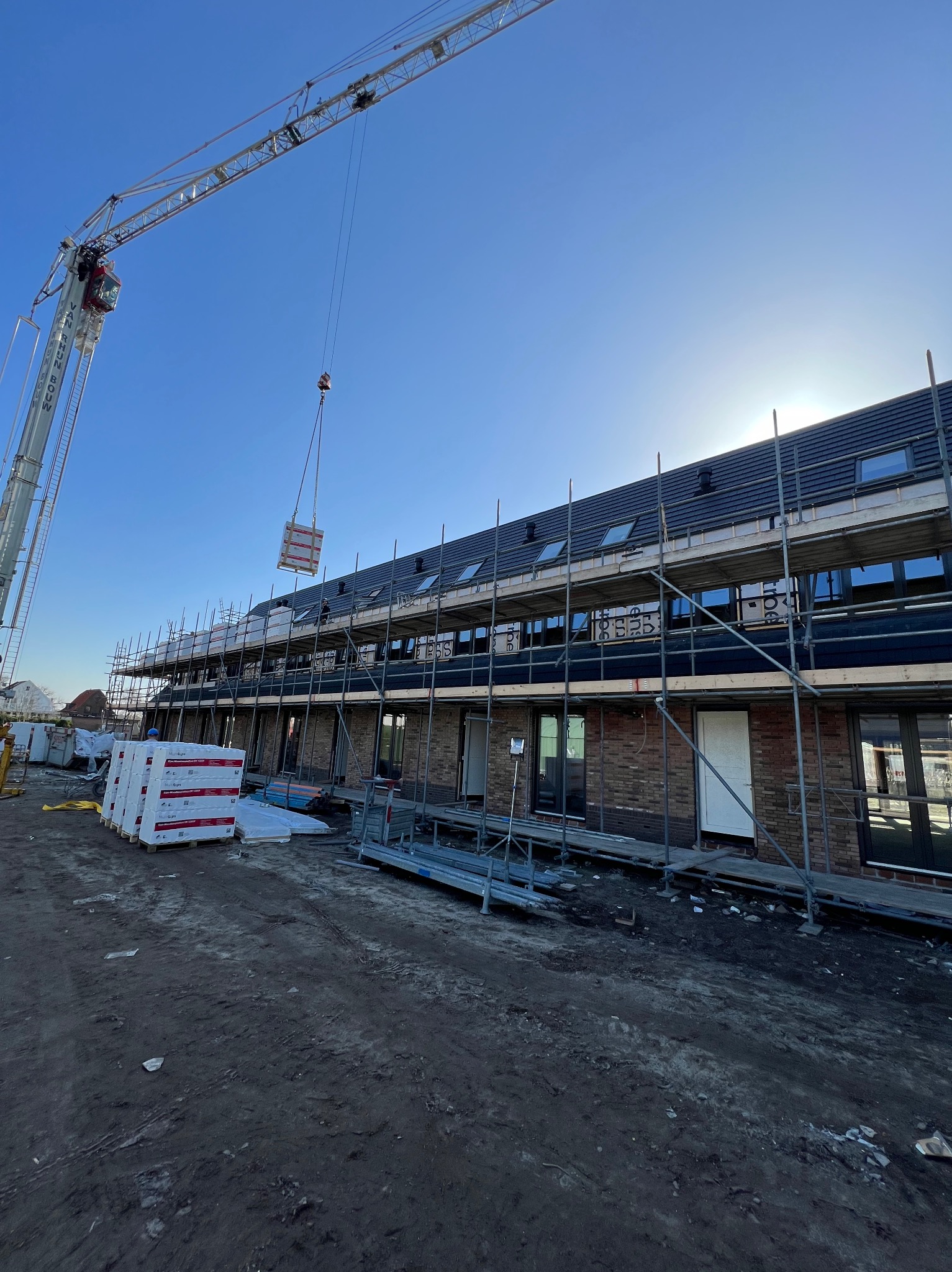 Montaż dachu na nowym budynku szeregowym; widoczne rusztowania, dźwig podnoszący materiały, palety z materiałami budowlanymi.