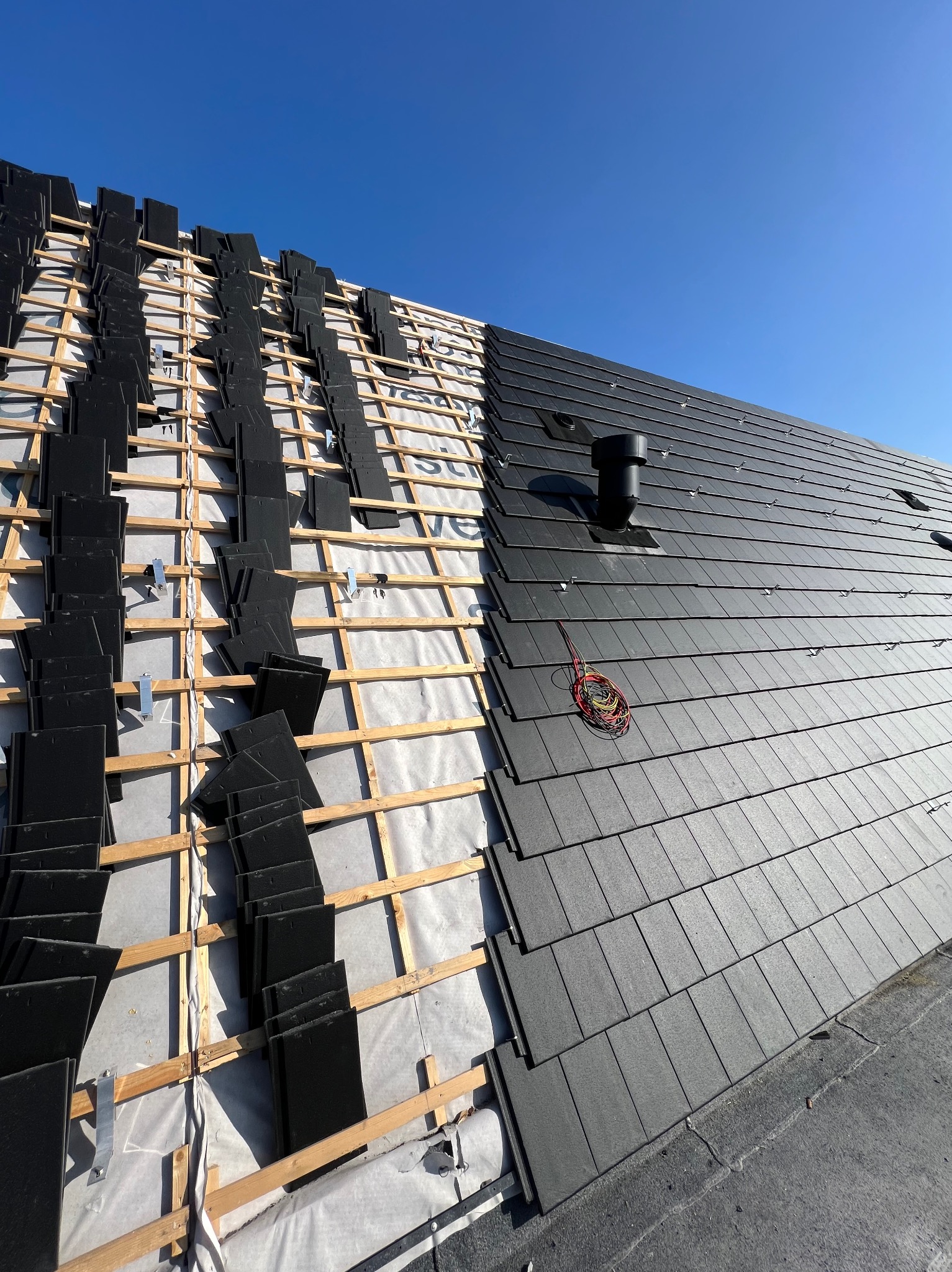 Częściowo pokryty dach z czarną dachówką, widoczna drewniana konstrukcja i nieułożone dachówki oraz przewody wentylacyjne na tle błękitnego nieba.