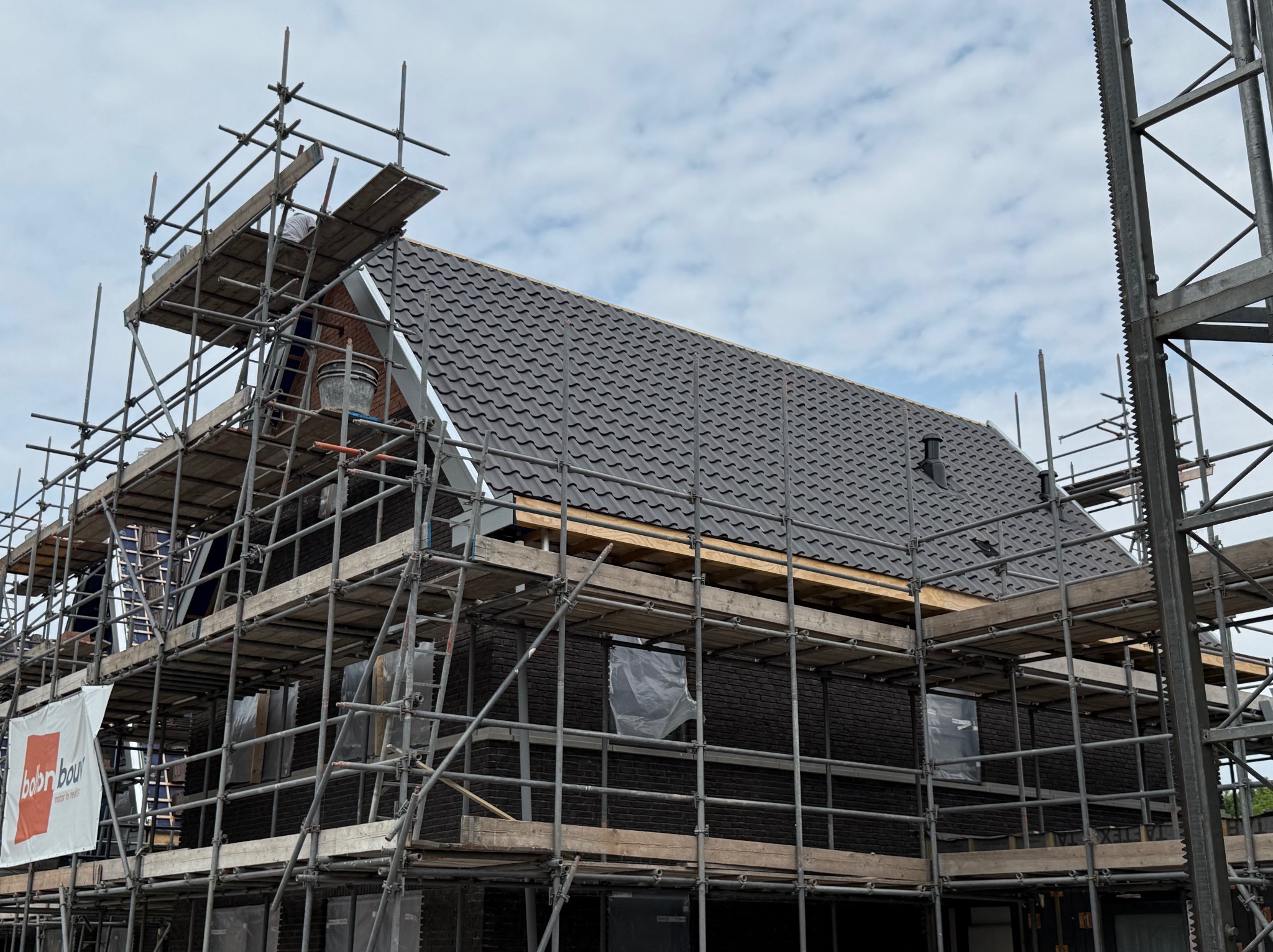 Dom w trakcie budowy z ciemną elewacją i szarym dachem, otoczony rusztowaniami. Widoczny fragment nieba z chmurami.