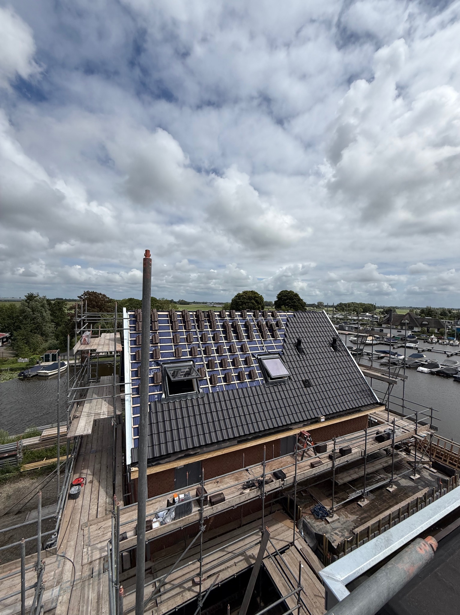 Częściowo pokryty dach z czarną dachówką, widoczne rusztowania, okna dachowe i elementy konstrukcji, z perspektywy z góry. W tle marina i krajobraz.