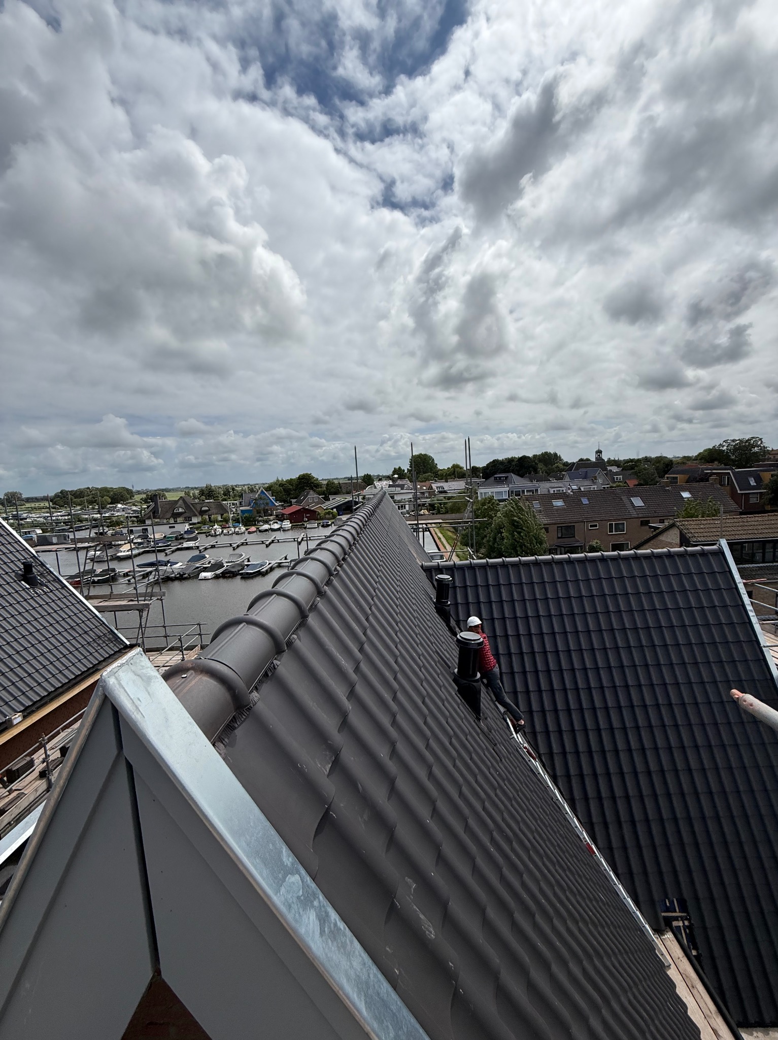 Ciemna dachówka na dachu domu z widokiem na miasto i pochmurne niebo. Dekarz w kasku na dachu. Widoczne obróbki blacharskie i elementy konstrukcji dachu.