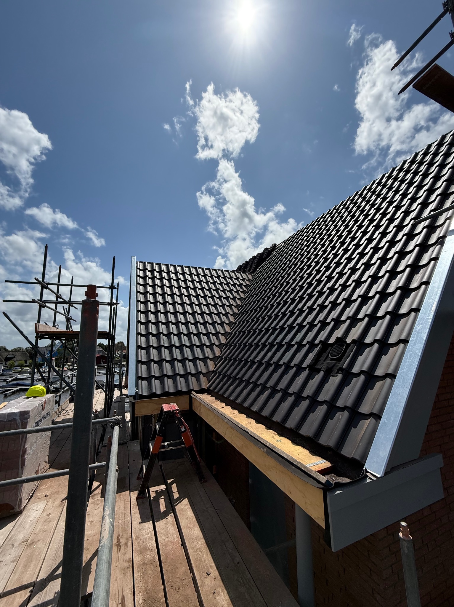 Czarne dachówki na połaci dachu w trakcie montażu, widok z rusztowania, słoneczny dzień, niebieskie niebo z chmurami.