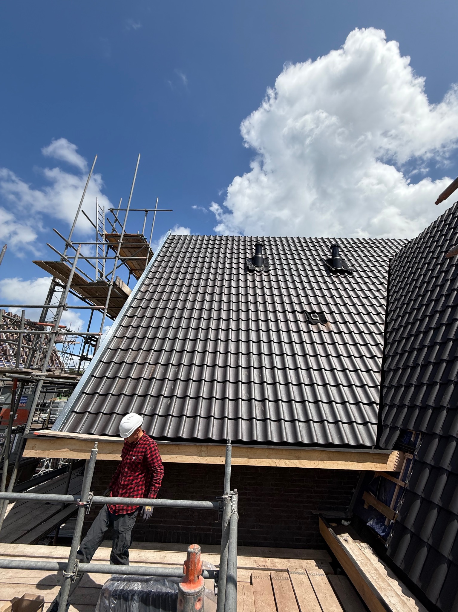 Prace dekarskie w Warszawie: Nowy dach z ciemnej dachówki ceramicznej, kominki wentylacyjne, widoczne rusztowania i dekarz w kasku podczas montażu.