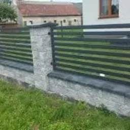 Nowoczesne ogrodzenie posesji z szarego kamienia i poziomych, czarnych, metalowych paneli, widoczne z perspektywy trawnika.