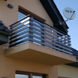 Nowoczesna balustrada balkonowa z poziomych, czarnych profili, zamontowana na balkonie domu jednorodzinnego z beżową elewacją, widok z dołu, na dachu antena satelitarna.