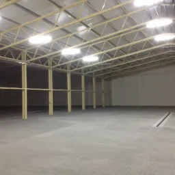 Wnętrze nowej, pustej hali z betonową posadzką, widoczne stalowe słupy i konstrukcja dachu z oświetleniem.