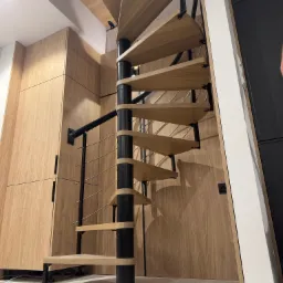 Spiralne schody z drewnianymi stopniami i czarną metalową konstrukcją, umieszczone w nowoczesnym wnętrzu z drewnianą zabudową.