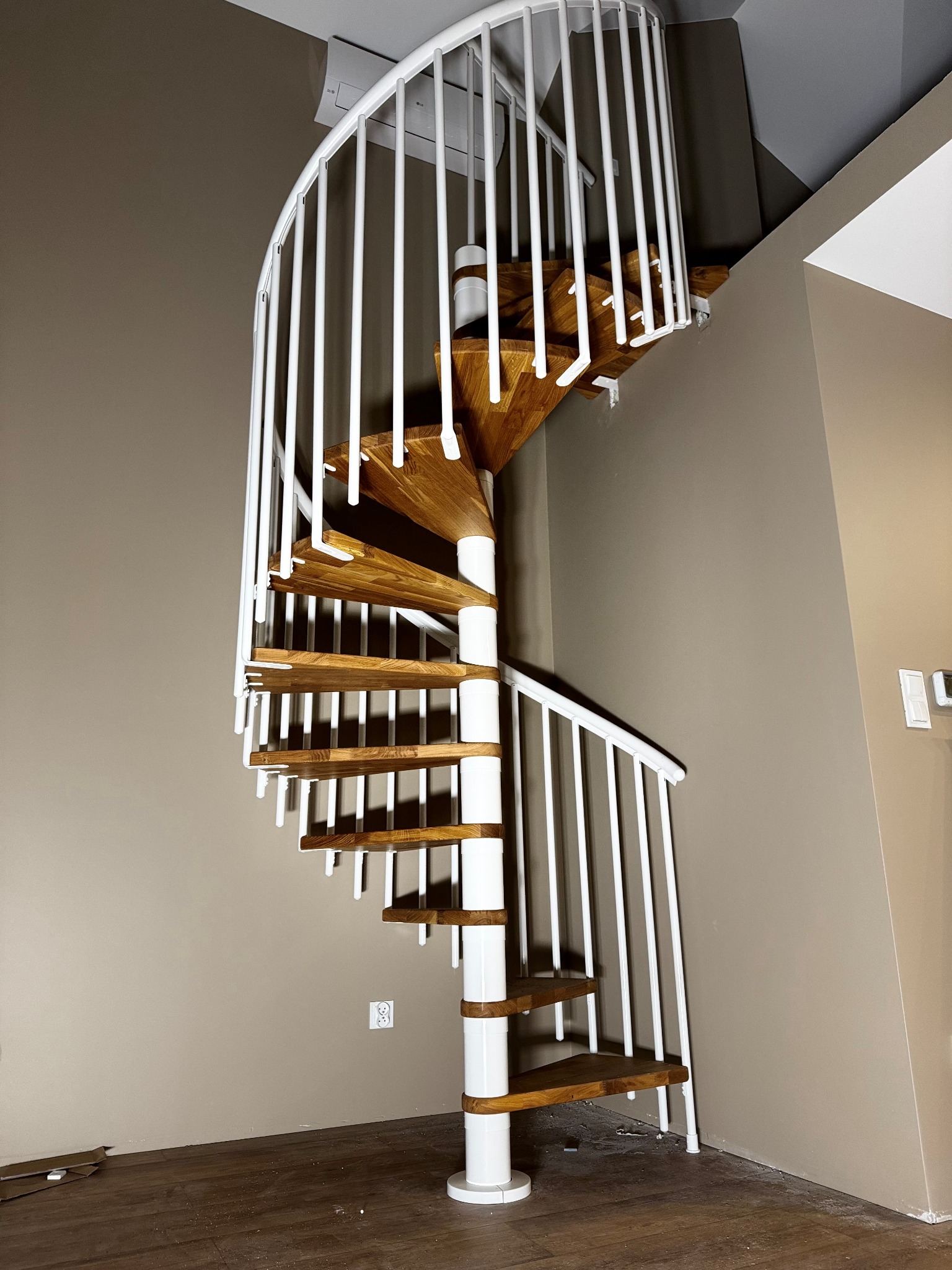 Spiralne schody z drewnianymi stopniami i białymi metalowymi barierkami, widoczne od dołu, w jasnym wnętrzu.