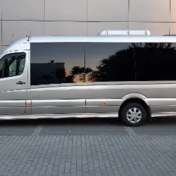 Srebrny bus pasażerski Mercedes-Benz Sprinter z przyciemnianymi szybami, zaparkowany na brukowanym parkingu przed budynkiem z szarej okładziny.