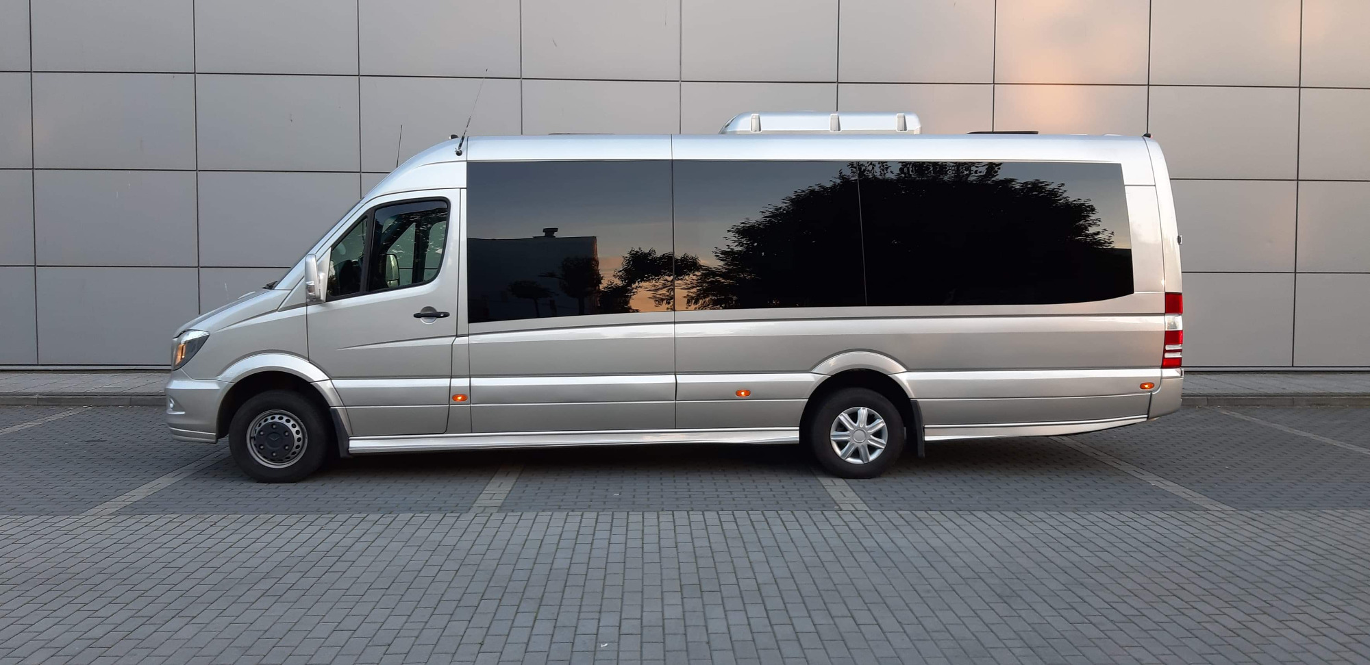 Srebrny bus pasażerski Mercedes-Benz Sprinter z przyciemnianymi szybami, zaparkowany na brukowanym parkingu przed budynkiem z szarej okładziny.