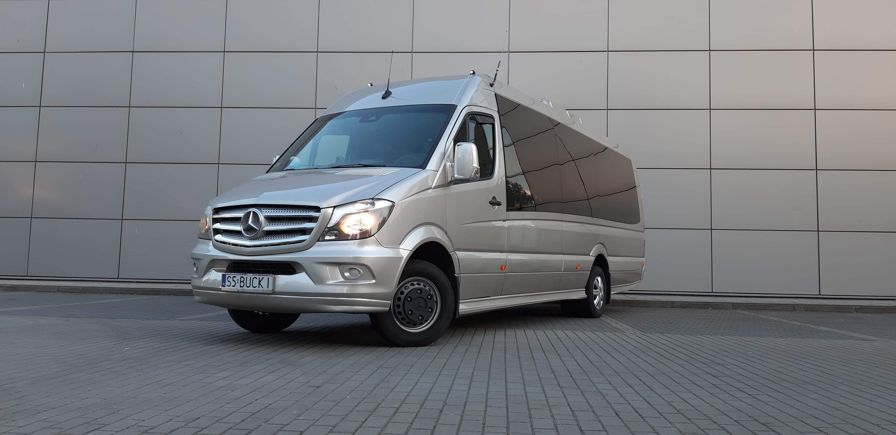 Srebrny bus Mercedes-Benz Sprinter z przyciemnianymi szybami na tle nowoczesnej ściany z paneli, widoczna tablica rejestracyjna SS-BUCK1.