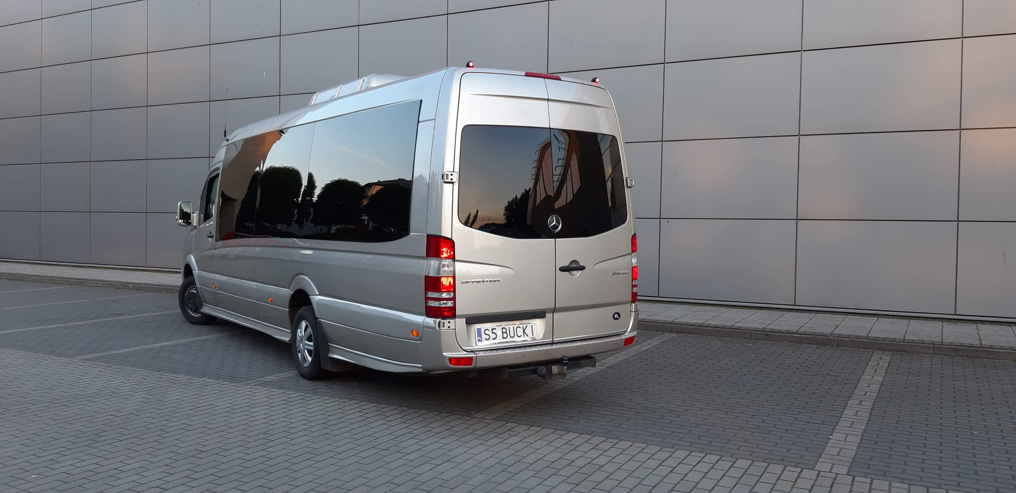Srebrny bus Mercedes Sprinter z przyciemnianymi szybami, zaparkowany na kostce brukowej przed nowoczesnym budynkiem z metalową elewacją. Tablica rejestracyjna 'S5 BUCK1'.