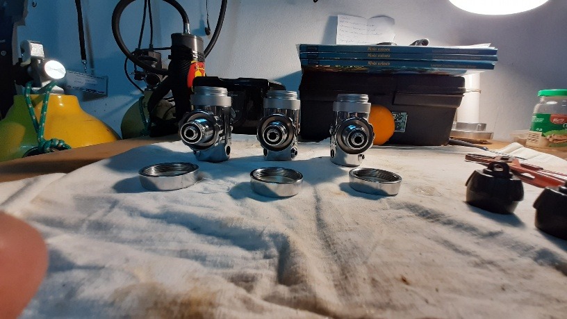 Trzy rozłożone automaty oddechowe do nurkowania, z odkręconymi pokrywami, leżące na białej szmatce na warsztatowym stole, w tle butla nurkowa, pomarańcza i książki.