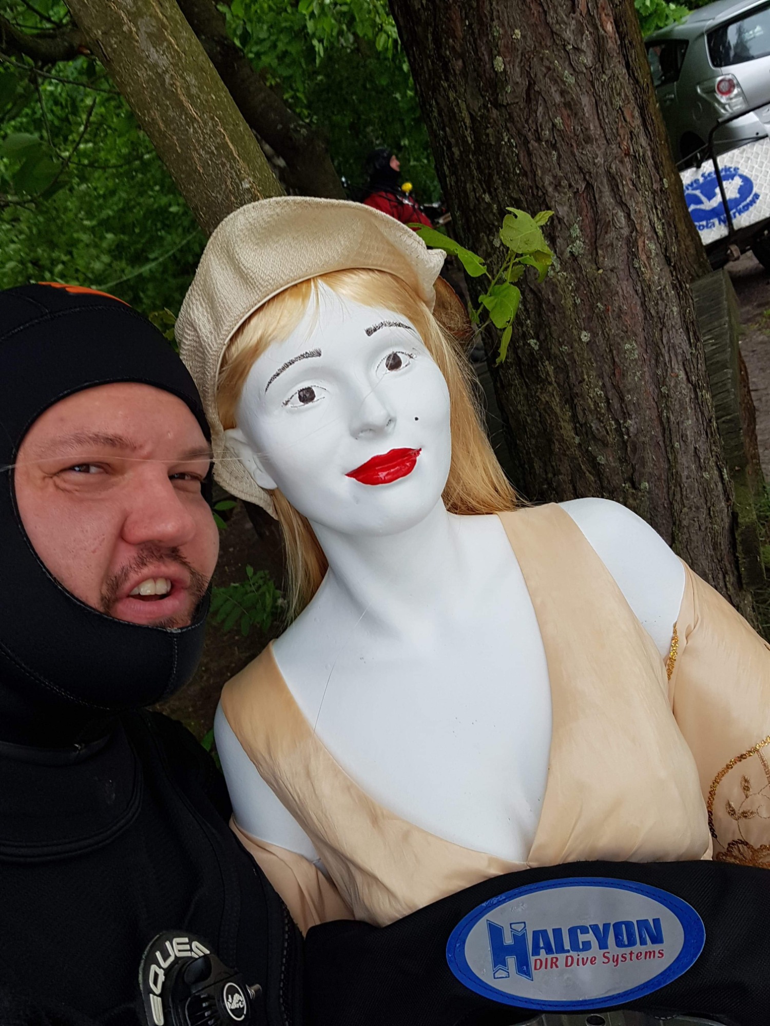 Mężczyzna w czarnym kombinezonie do nurkowania robi selfie z manekinem ubranym w beżową sukienkę i kapelusz przy drzewie, widoczny napis Halcyon DIR Dive Systems.
