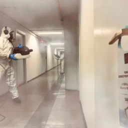 Dezynfekcja korytarza biurowego przez dw&oacute;ch pracownik&oacute;w w kombinezonach ochronnych, z użyciem zamgławiacza, widoczna zawieszka na drzwiach z napisem 'Obiekt zdezynfekowany'.