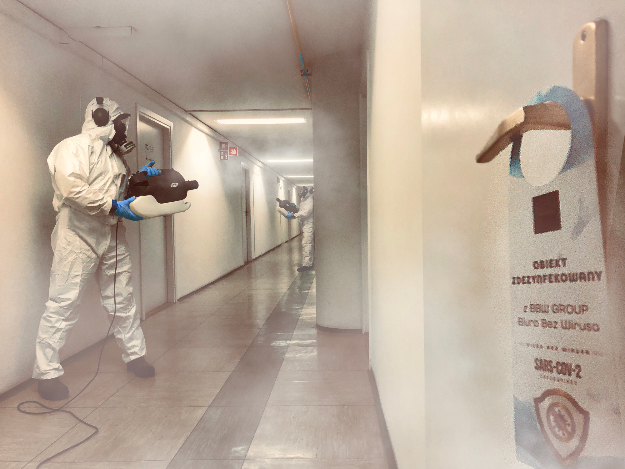 Dezynfekcja korytarza biurowego przez dwóch pracowników w kombinezonach ochronnych, z użyciem zamgławiacza, widoczna zawieszka na drzwiach z napisem 'Obiekt zdezynfekowany'.