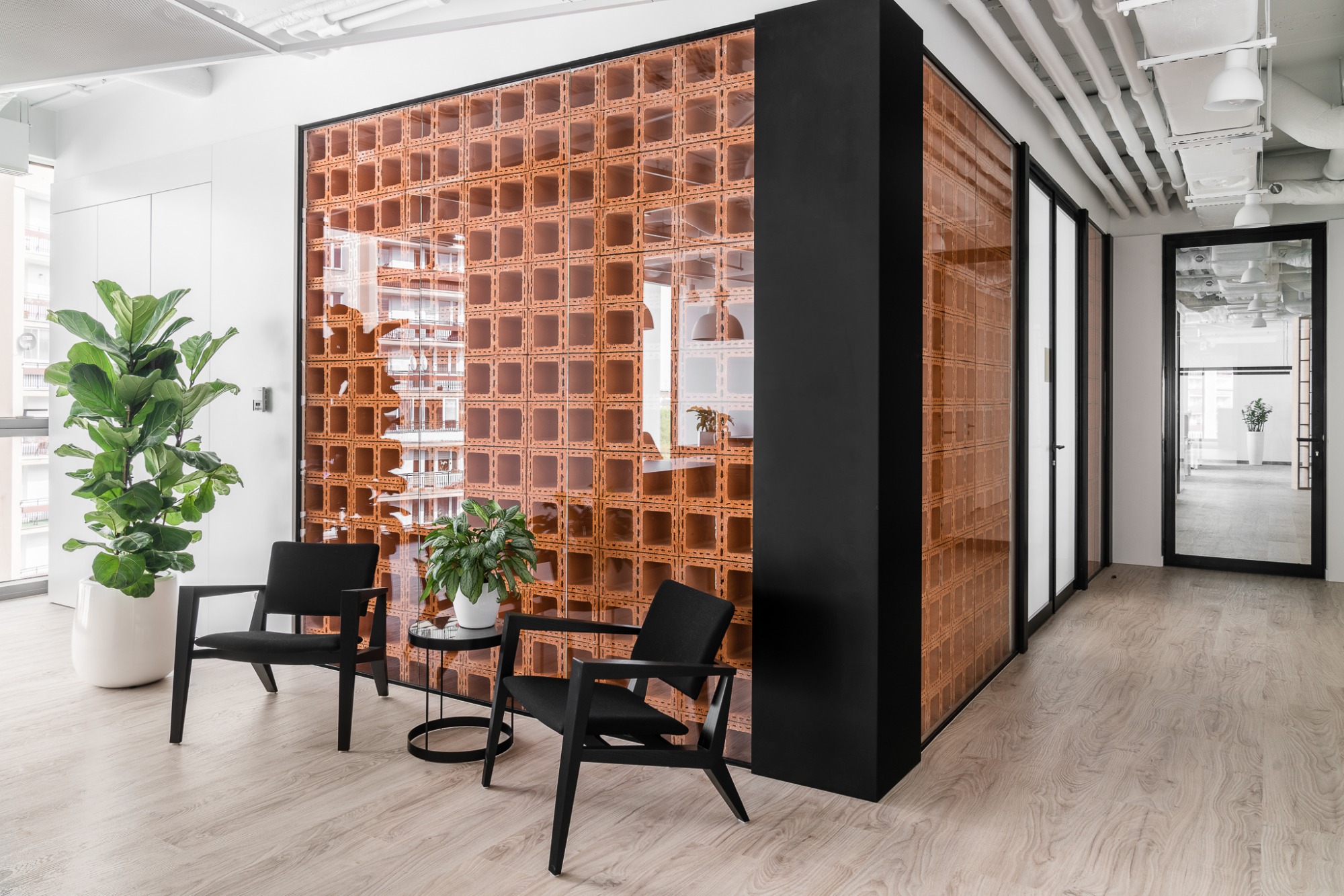 Nowoczesne biuro z dekoracyjną ścianą z ażurowych, terakotowych bloczków, uzupełnione minimalistycznymi czarnymi krzesłami i roślinami doniczkowymi, podłoga wyłożona panelami imitującymi jasne drewno.