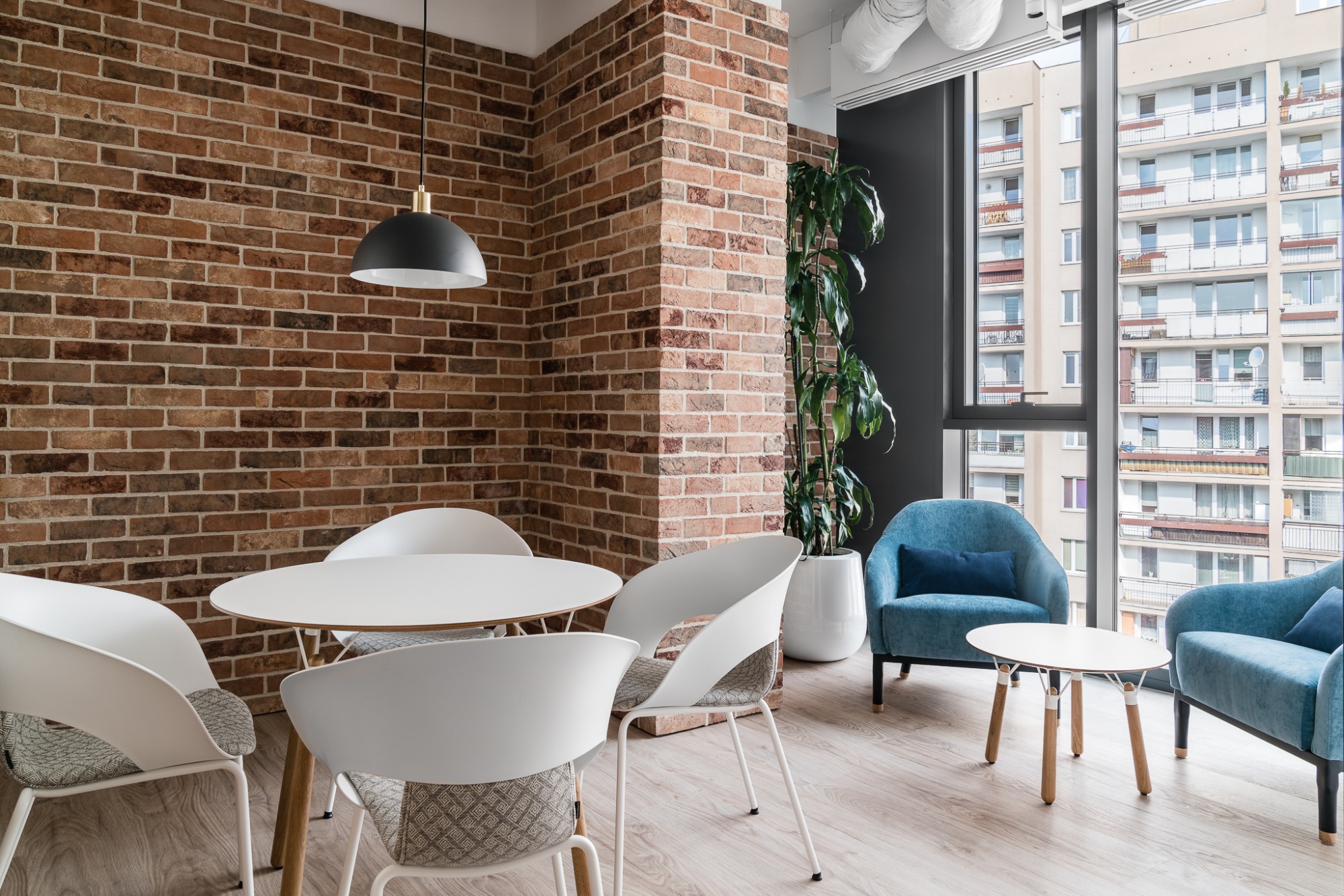 Jasne biuro z ceglaną ścianą, białymi krzesłami i stołem, błękitnymi fotelami, dużym oknem z widokiem na budynki mieszkalne oraz rośliną doniczkową.