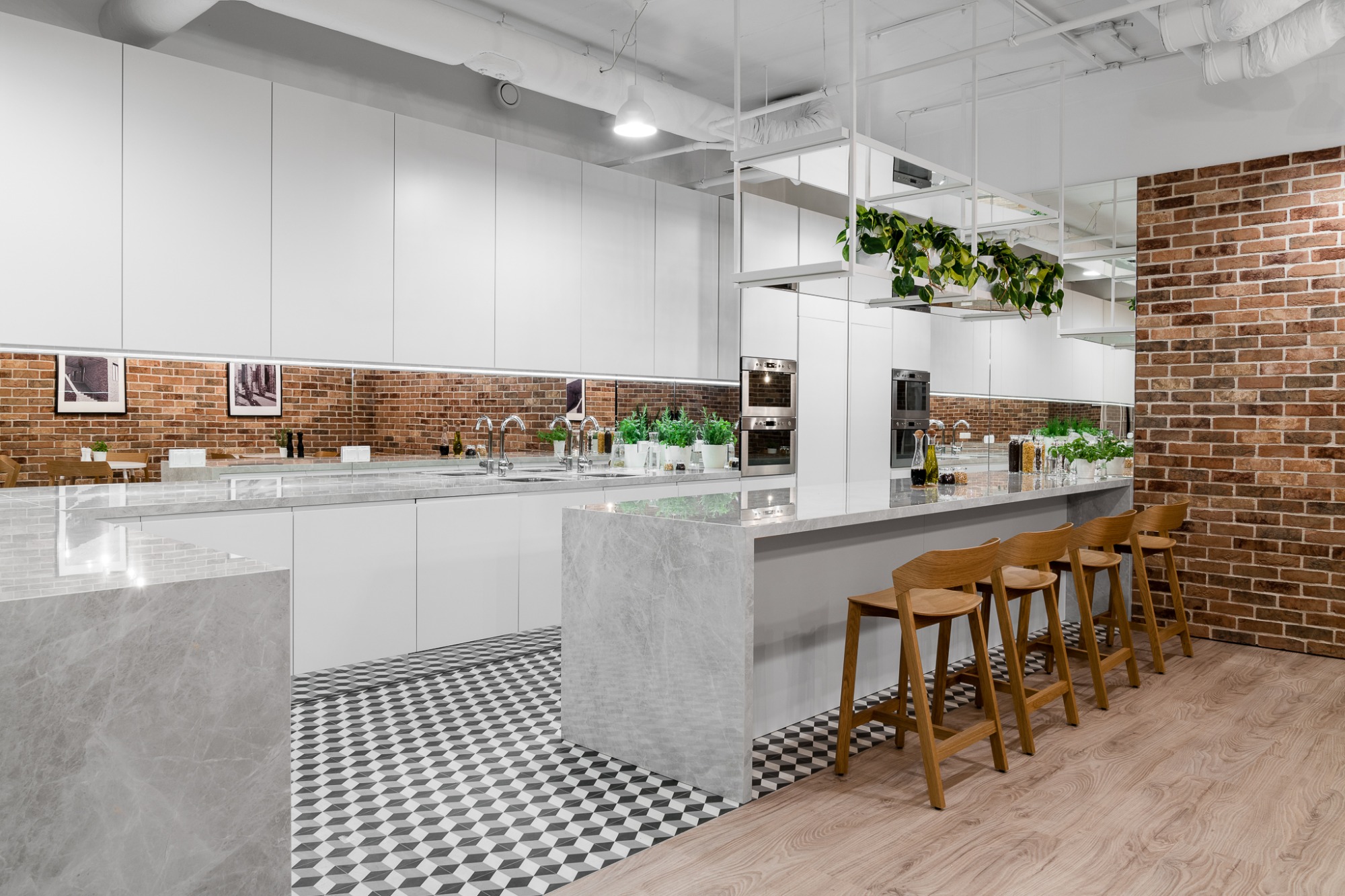Nowoczesna kuchnia biurowa z białymi szafkami, marmurowym blatem, ceglaną ścianą w tle i geometryczną podłogą, uzupełniona drewnianymi stołkami barowymi i wiszącymi roślinami.