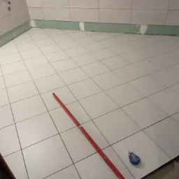 Białe płytki podłogowe w trakcie układania w narożnej łazience, widoczna poziomica i zwijana miarka, ściany częściowo przygotowane do dalszych prac wykończeniowych.