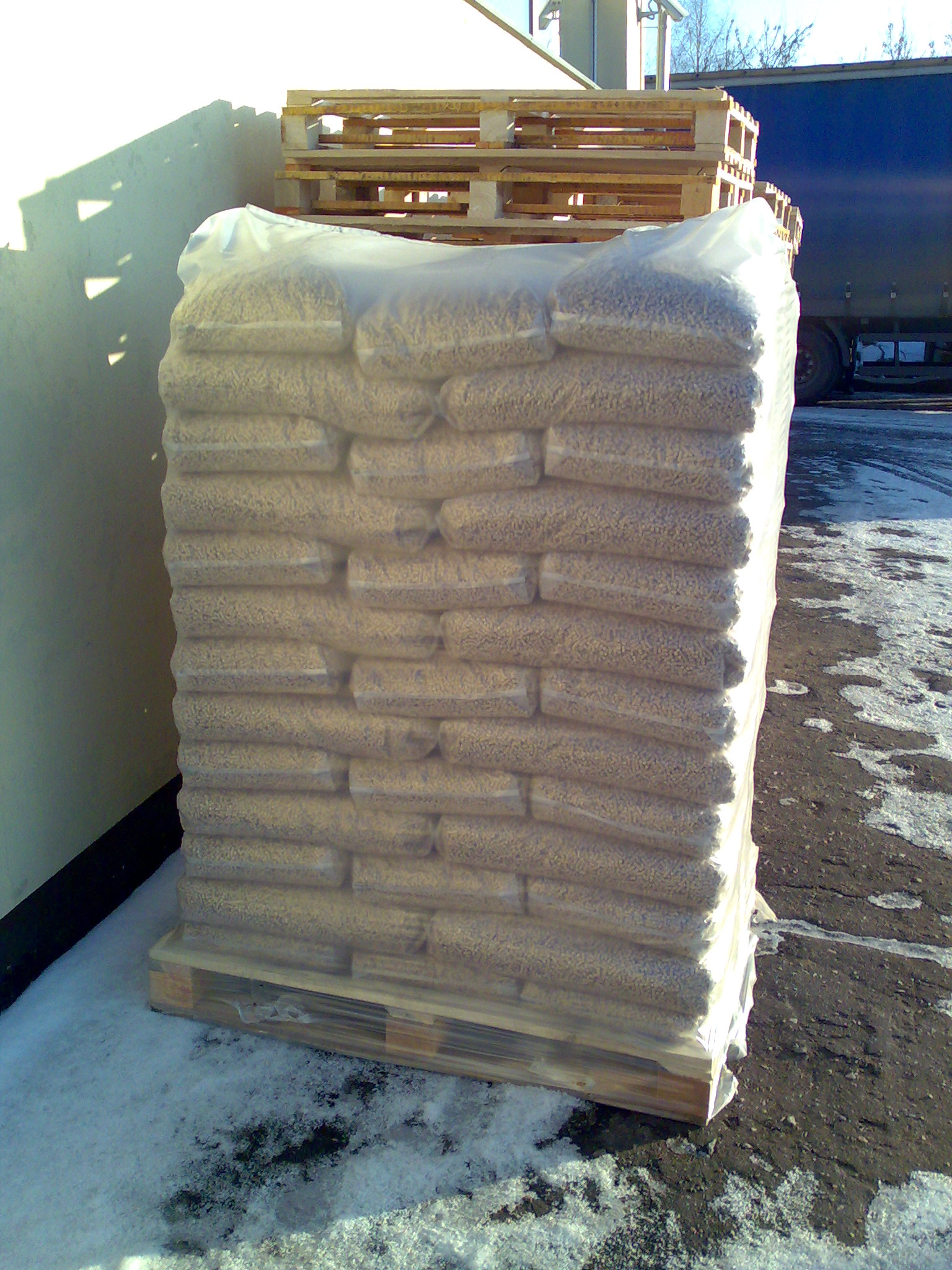 Ustawiony na palecie wysoki stos worków z granulowanym materiałem opałowym, owinięty przezroczystą folią, na zewnątrz, obok ściany budynku i zaparkowanej ciężarówki, z resztkami śniegu na ziemi.
