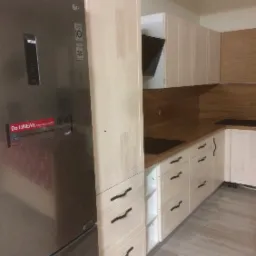 Jasna zabudowa kuchenna z drewnianym blatem, widoczny fragment lodówki LG z dystrybutorem wody, minimalistyczne czarne uchwyty szuflad.