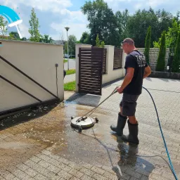 Pracownik w gumowych butach czyści kostkę brukową na posesji za pomocą okrągłej myjki ciśnieniowej, widoczne zabrudzenia i woda.