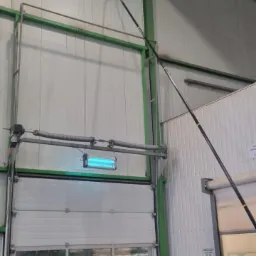 Czarny kij teleskopowy z końcówką czyszczącą sięga pod sufit hali przemysłowej, widoczna brama segmentowa i konstrukcja stalowa budynku.