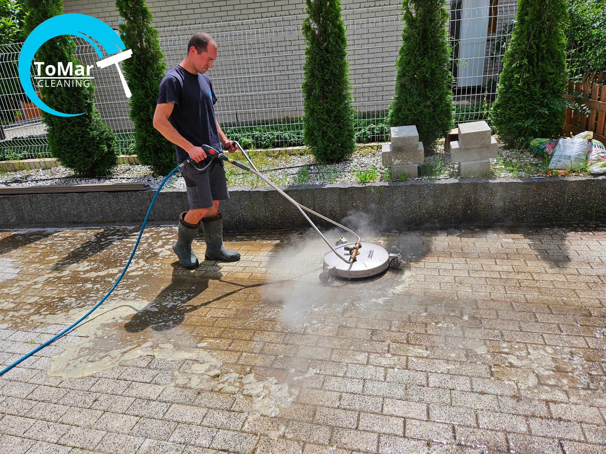 Mężczyzna w gumowych butach czyści kostkę brukową za pomocą myjki ciśnieniowej z okrągłą dyszą; widoczny efekt czyszczenia i logo firmy w lewym górnym rogu.