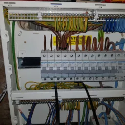 Otwarta skrzynka rozdzielcza z widocznymi bezpiecznikami, przewodami o różnych kolorach (niebieski, brązowy, żółto-zielony) i listwami zaciskowymi, z naklejką ostrzegawczą.