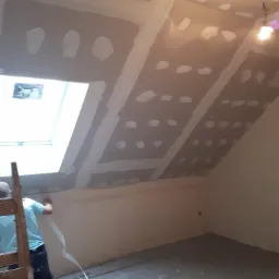 Poddasze w trakcie prac wykończeniowych: montaż płyt kartonowo-gipsowych, widoczne łączenia wzmocnione taśmą, pracownik na drabinie przy oknie dachowym.