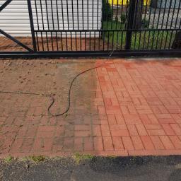 Mr.D Usługi Specjalistyczne - Częściowo wyczyszczona, czerwona kostka brukowa przed czarną metalową bramą, widoczna różnica w kolorze i czystości między umytą a nieumytą powierzchnią, leżący wąż ciśnieniowy.