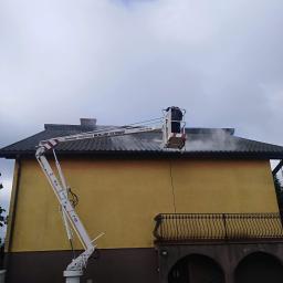 Mr.D Usługi Specjalistyczne - Czyszczenie dachu z drabiny koszowej, widoczny strumień wody pod ciśnieniem usuwający zabrudzenia z dachówki, budynek z balkonem i żółtą elewacją w tle.