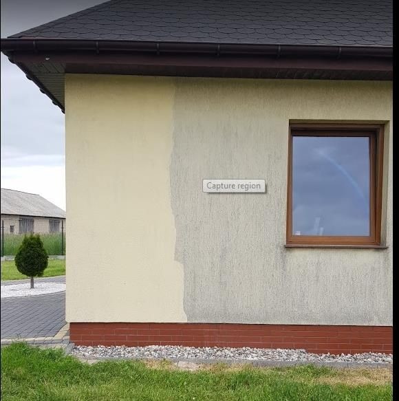 Fragment elewacji budynku z widoczną różnicą w kolorze po częściowym czyszczeniu, brudna i czysta powierzchnia obok siebie, drewniane okno, ceglana podmurówka.