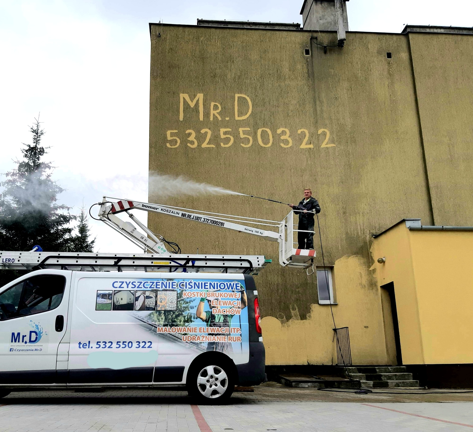 Pracownik na podnośniku koszowym czyści elewację budynku z napisem 'Mr.D 532550322' za pomocą myjki ciśnieniowej, w tle widoczny samochód dostawczy z reklamą firmy.