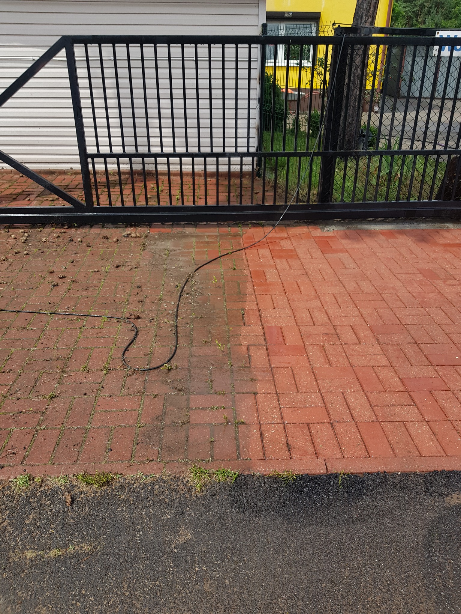Częściowo wyczyszczona, czerwona kostka brukowa przed czarną metalową bramą, widoczna różnica w kolorze i czystości między umytą a nieumytą powierzchnią, leżący wąż ciśnieniowy.