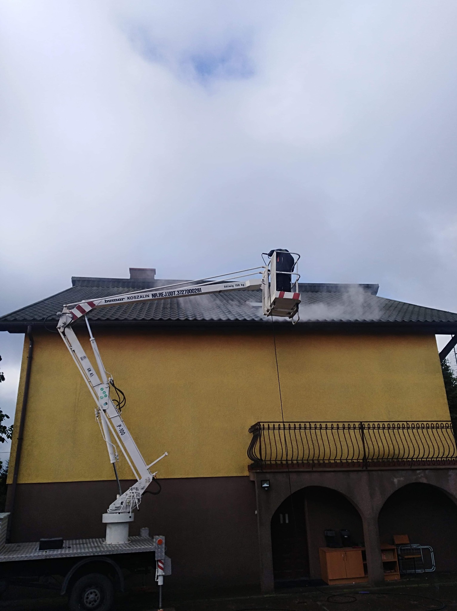 Czyszczenie dachu z drabiny koszowej, widoczny strumień wody pod ciśnieniem usuwający zabrudzenia z dachówki, budynek z balkonem i żółtą elewacją w tle.