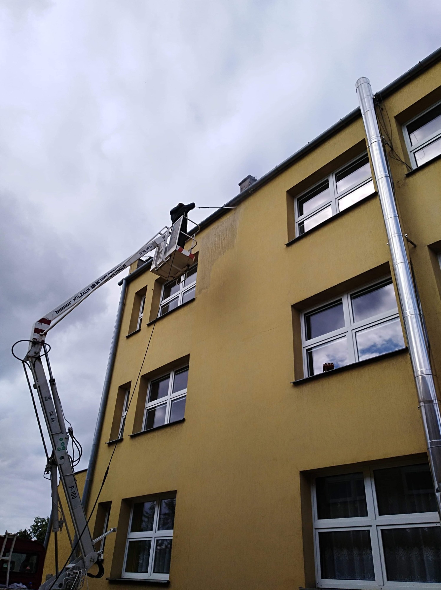 Pracownik na podnośniku koszowym myje elewację budynku, widoczne ślady czyszczenia na żółtej ścianie, metalowy komin odprowadzający spaliny na zewnątrz.