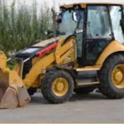 Żółta koparko-ładowarka Caterpillar z opuszczoną łyżką i podporą, zaparkowana na asfaltowej powierzchni na tle zieleni.