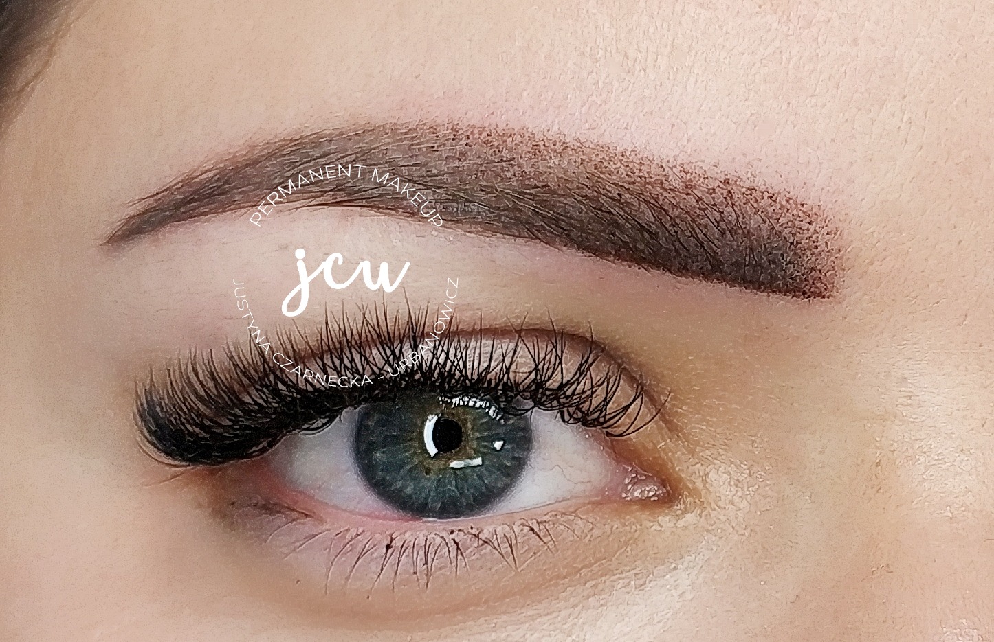 Zbliżenie na oko z makijażem permanentnym brwi metodą pudrową i przedłużonymi rzęsami, widoczny delikatny cień do powiek i logo firmy.