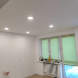 Wnętrze pokoju po remoncie w Ostrołęce z nowym sufitem podwieszanym z oświetleniem punktowym, świeżo pomalowanymi ścianami i ułożonymi panelami podłogowymi. Widoczne przygotowane przyłącza...