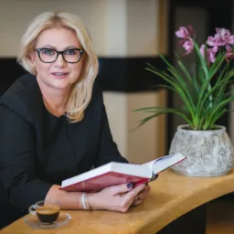 Kobieta w czarnej marynarce i okularach siedzi przy marmurowym stole, trzyma otwartą książkę z czerwoną okładką, obok filiżanka espresso i doniczka z storczykami.