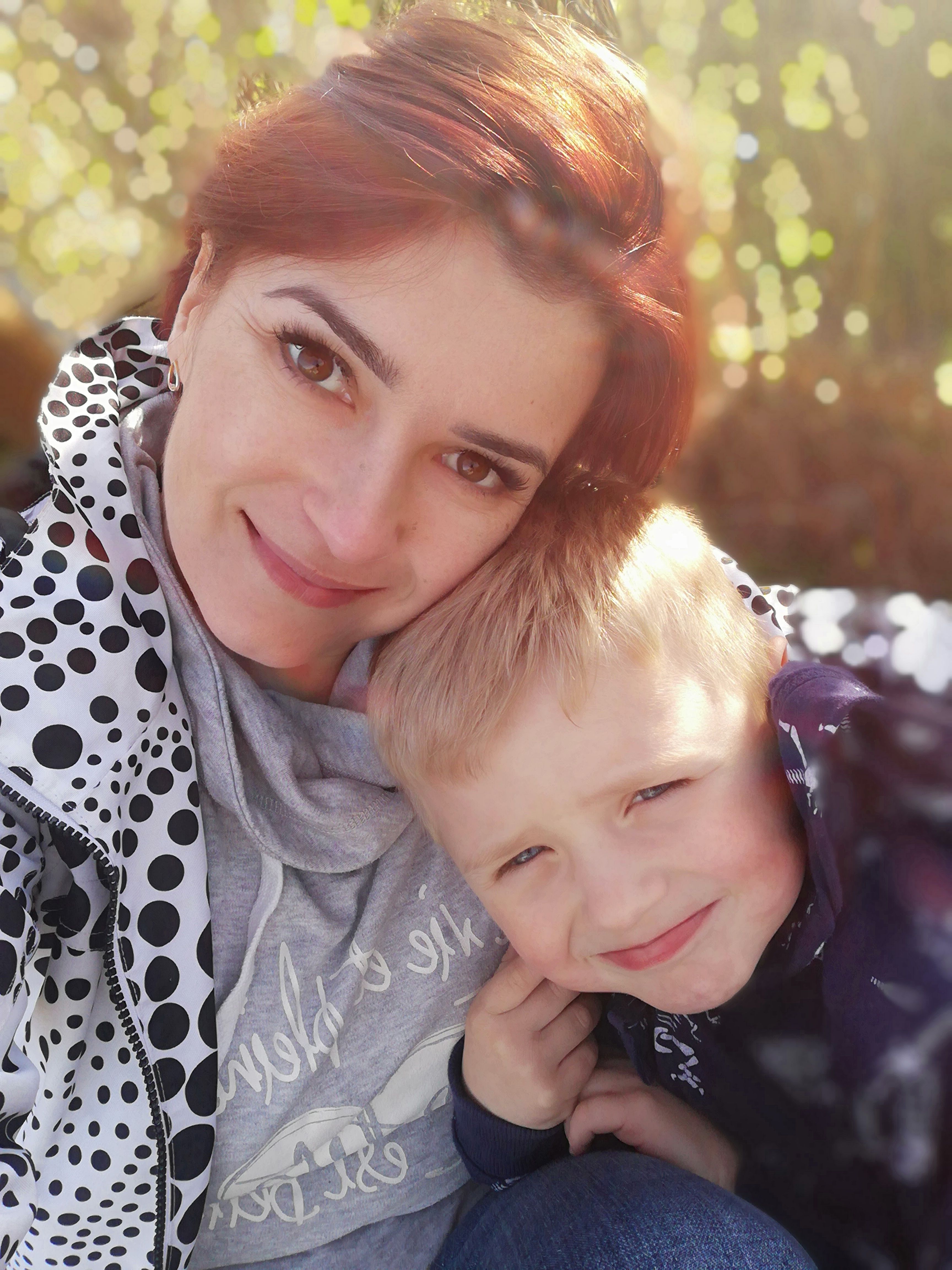 Uśmiechnięta kobieta z rudymi włosami przytula blond chłopca na tle rozmytych drzew i słonecznego światła.