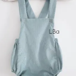 Dziecięcy rampers z błękitnego sztruksu, wiszący na białym wieszaku z motywem misia, widoczne metalowe zatrzaski i gumki ściągające przy nogawkach.